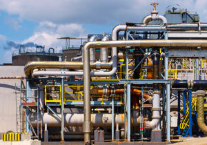 An industrial facility with a complex network of pipelines and machinery, featuring large metal pipes, valves, and tanks under a partly cloudy sky.