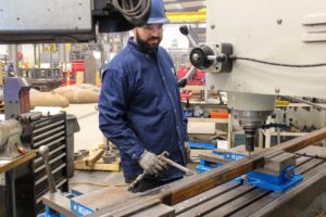 Worker in blue safety gear operating a large industrial drilling machine in a factory workshop.