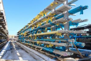 Rows of metal pipes covered in protective plastic wrap stored on outdoor shelving under a clear blue sky.