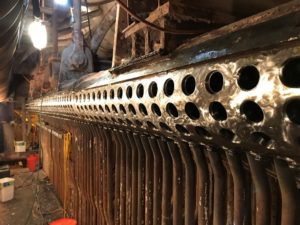 Close-up of an industrial boiler’s tubes and manifold with multiple circular openings, showing intricate metalwork and various components.