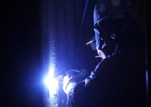 A welder in protective gear, including a helmet and mask, is performing a welding task, with bright blue sparks illuminating the scene.