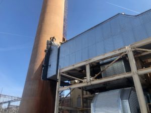 Large industrial chimney and steel structure with corrugated metal siding, set against a clear blue sky with a visible contrail.