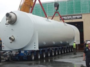 A large white industrial pressure vessel is being lifted by a crane outside Helfrich Brothers Boiler Works, Inc., with workers nearby.
