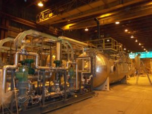 A large industrial pressure vessel with interconnected piping and valves inside a spacious, well-lit manufacturing facility.