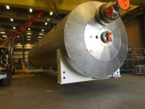 A large, cylindrical pressure vessel being lifted by a crane inside an industrial facility, with a red truck visible nearby.