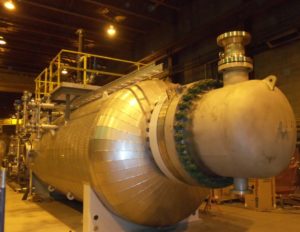 A large, cylindrical industrial pressure vessel with a rounded end cap and multiple connections sits in a well-lit factory setting.