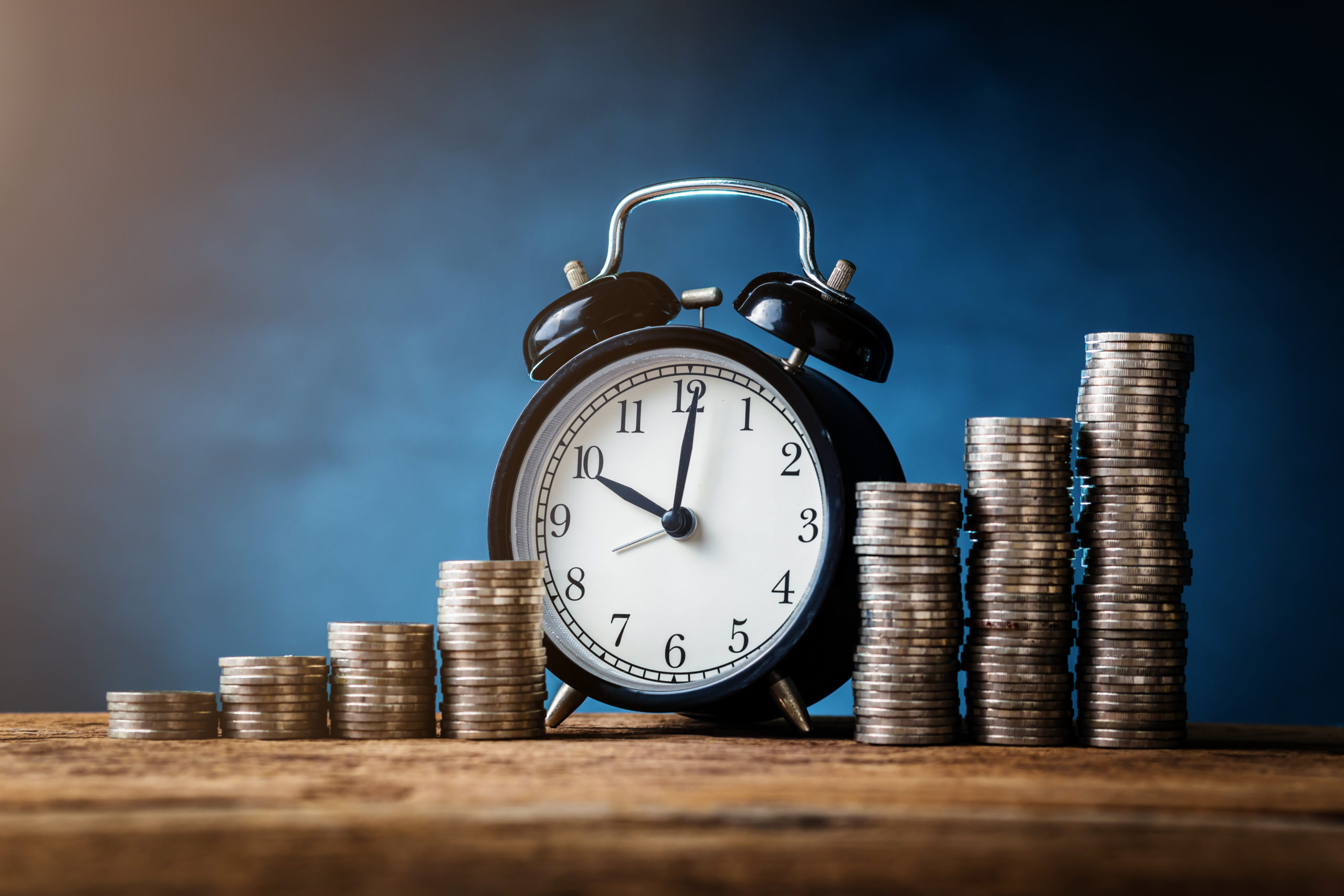 shutterstock_706427383 An old-fashioned alarm clock next to stacks of coins arranged in ascending order on a wooden surface, symbolizing time and money.