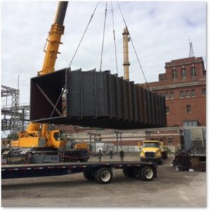 A large metal structure being hoisted by a crane onto a flatbed truck at an industrial site with brick buildings.