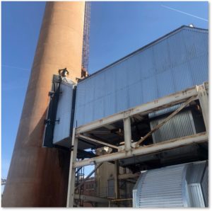 View of an industrial plant with a tall, rust-colored smokestack and large metal structures under a clear blue sky.