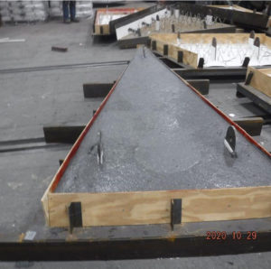 Concrete slab curing in a triangular wooden form with metal hooks, surrounded by other forms in an industrial setting.