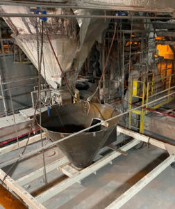 A large industrial funnel being installed in a factory, supported by chains and metal framing, with machinery nearby.