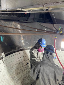 Two workers in protective suits and helmets performing maintenance inside a large industrial unit with white tiles.