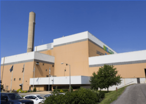 A large industrial building with beige walls and a tall chimney is surrounded by flags and greenery, with a clear blue sky overhead.