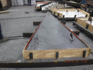 A triangular metal part being prepped for installation, secured in a wooden frame with attachment points.