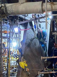 A worker in protective gear operates near a suspended metal component, surrounded by scaffolding, pipes, and bright lights.