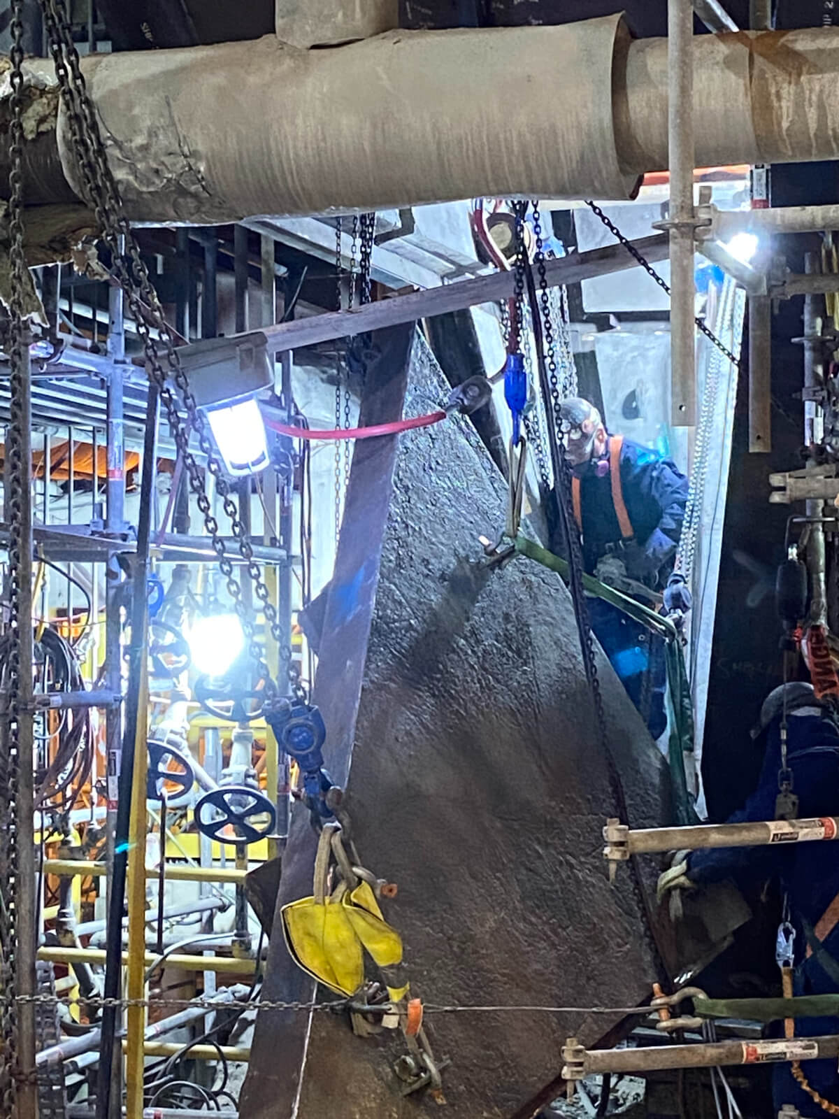 IMG_0792 A worker in protective gear operates near a suspended metal component, surrounded by scaffolding, pipes, and bright lights.