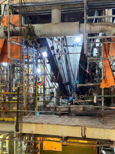 A construction or maintenance area inside an industrial facility with scaffolding, pipes, and chains. Bright lights illuminate the workspace, and orange tarps provide barriers or cover for ongoing work.