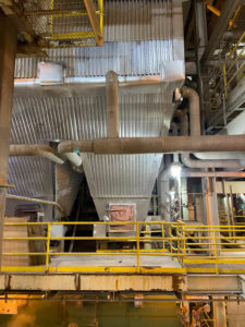 Interior view of an industrial facility showing large corrugated metal surfaces, pipes, and yellow guardrails surrounding the equipment.