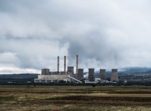 A large industrial power plant with multiple cooling towers emitting steam, set against a cloudy sky in a rural landscape.