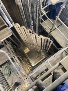 A worker looking down a scaffolded industrial shaft as a large heat exchanger coil is being lifted out during maintenance.