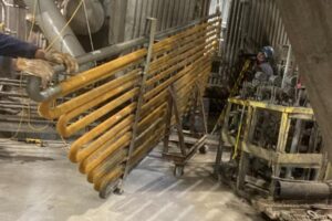 Worker handling a large rusted heat exchanger coil inside an industrial facility during maintenance work.